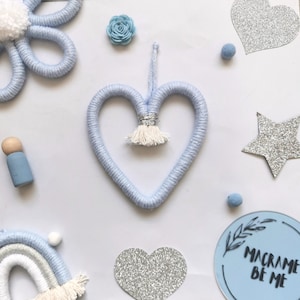 May include: A light blue macrame heart with a silver and white tassel, surrounded by decorative elements. Includes a blue flower, a wooden peg doll, a rainbow, and glittery hearts and stars. The text "MACRAME BE ME" is also visible.