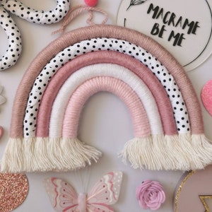 May include: A pink, white and brown macrame rainbow wall hanging with white fringe. The rainbow is made of yarn and has a black and white polka dot pattern on the top layer. The sign above the rainbow reads "Macrame Be Me."