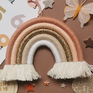 May include: A handcrafted macrame rainbow in peach, gold, beige, and white. The rainbow features a glittery gold centre and fringe at the base. Decorative stars and a butterfly are in the background.