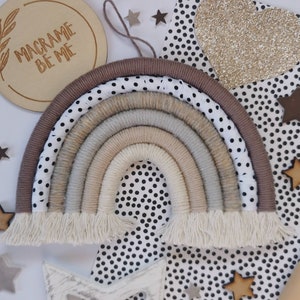 May include: A macrame rainbow wall hanging with a brown, beige, and white color scheme. The rainbow is made of yarn and has a fringe at the bottom. The rainbow is hanging on a black and white polka dot background.