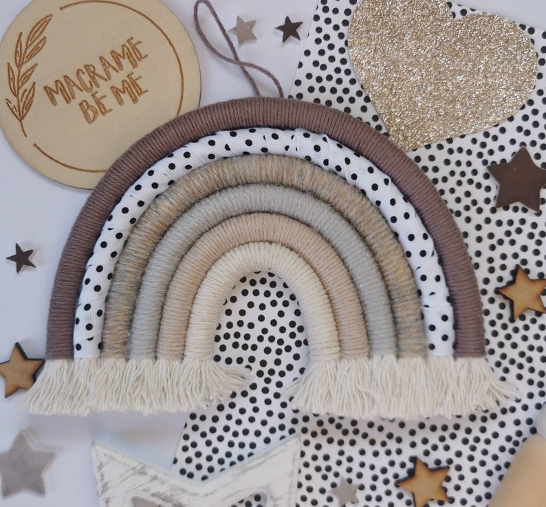May include: A macrame rainbow wall hanging with a brown, beige, and white color scheme. The rainbow is made of yarn and has a fringe at the bottom. The rainbow is hanging on a black and white polka dot background.
