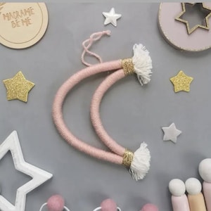 May include: A pink macrame crescent moon with gold detailing and white tassels. The image also features gold and white star decorations, and a wooden disc with the text "MACRAME BE ME".