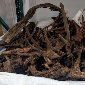 May include: A large white bag filled with various pieces of driftwood. The wood pieces are primarily brown with a range of textures and shapes, including branches and root structures. The bag is open, revealing the contents.