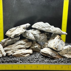 May include: A collection of light gray and tan aquarium rocks arranged on a bed of black gravel. The rocks have a layered texture and varying sizes. A yellow ruler is in the foreground.