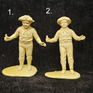 May include: Two plastic figurines of boys wearing scout uniforms and hats. The boys are standing on separate bases and are holding their right hands up in a salute. The figurines are beige in color.