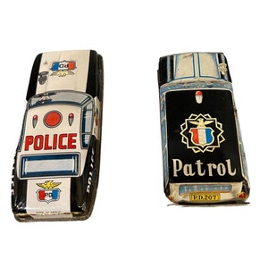 May include: Two vintage toy police cars. One is white and black with "POLICE" printed on the side. The other is black with "Patrol" printed on the side. Both have a retro design and are made of metal.