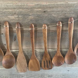 May include: A set of eight brown wooden kitchen utensils hanging from a thin string. The collection includes various spoons, a flat spatula, a slotted turner, a perforated spoon, and a salad fork, all featuring a natural wood grain finish.