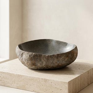 May include: A dark gray stone bowl with a rough, textured exterior and a smooth, polished interior basin. The vessel features an irregular, organic shape with a thick rim and natural variations in the stone surface color and texture.