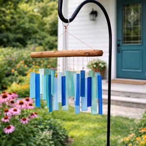 May include: A wind chime featuring a horizontal piece of natural driftwood suspended from a black metal hook. Multiple rectangular glass pieces in varying shades of blue, teal, and light aqua are attached to the wood with thin copper-colored wires.