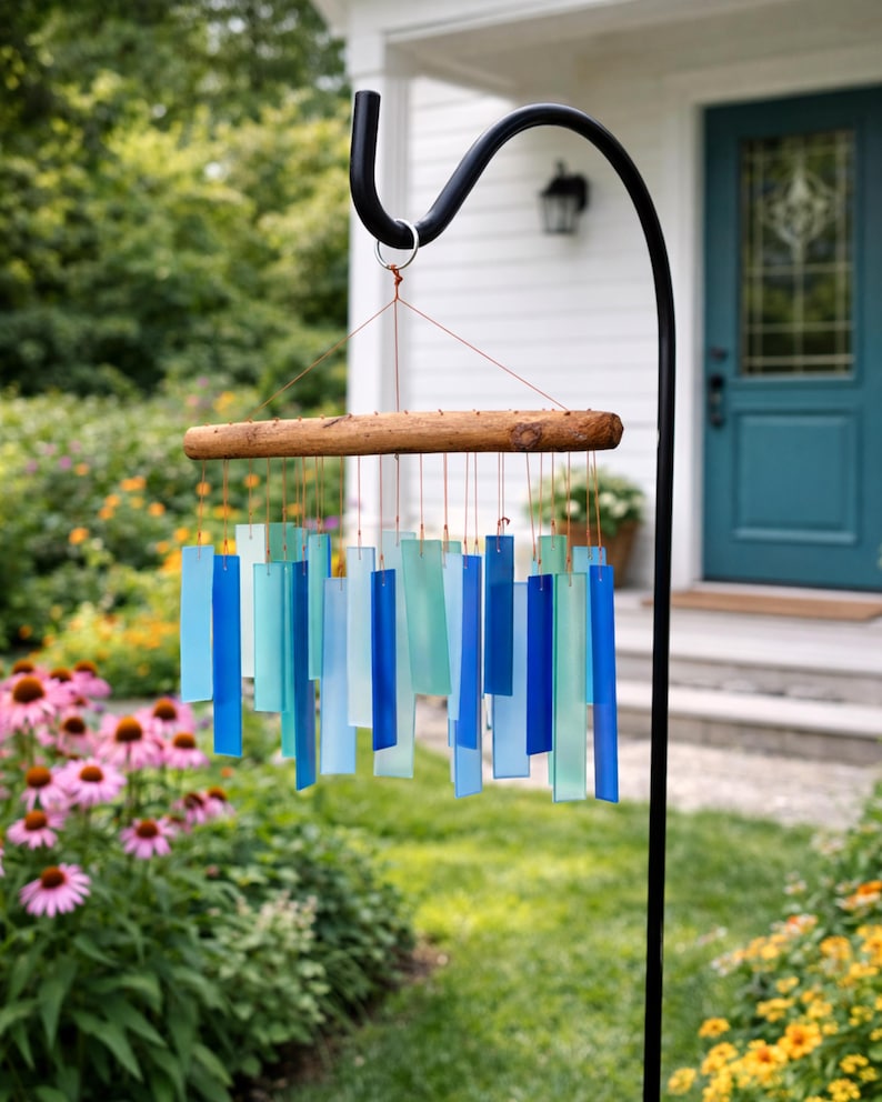 May include: A wind chime featuring a horizontal piece of natural driftwood suspended from a black metal hook. Multiple rectangular glass pieces in varying shades of blue, teal, and light aqua are attached to the wood with thin copper-colored wires.