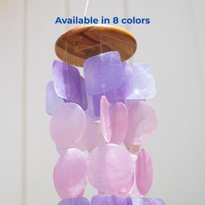 May include: A hanging wind chime with a wooden top and strands of purple and pink shell-shaped pieces. The text "Available in 8 colors" is displayed in blue. The chime is designed for outdoor or indoor decor.