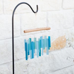 May include: A wind chime featuring multiple rectangular, translucent, and opaque blue and teal acrylic strips suspended from a natural light-colored wooden branch by thin metallic wires, hanging from a black metal shepherd's hook.