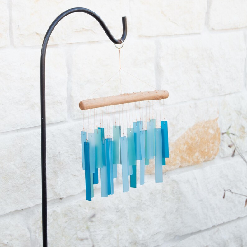 May include: A wind chime featuring multiple rectangular, translucent, and opaque blue and teal acrylic strips suspended from a natural light-colored wooden branch by thin metallic wires, hanging from a black metal shepherd's hook.