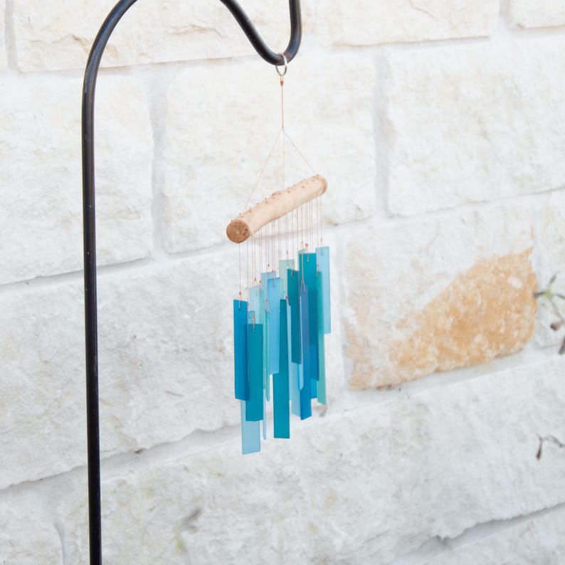 May include: A wind chime featuring multiple rectangular glass pieces in varying shades of blue and teal, suspended by thin copper wires from a horizontal piece of light-colored natural wood, hanging from a black metal shepherd's hook.
