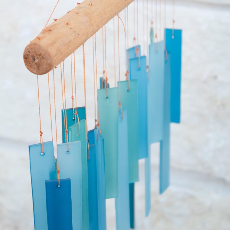 May include: A wind chime featuring multiple rectangular glass panels in various shades of blue, ranging from light cyan to deep navy. The glass pieces are suspended by thin orange strings from a cylindrical wooden branch at the top.
