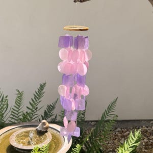 May include: A hanging wind chime with a wooden top and strands of pink and purple shell-like pieces. The wind chime is suspended from a small branch against a neutral background. The design is decorative and suitable for outdoor use.