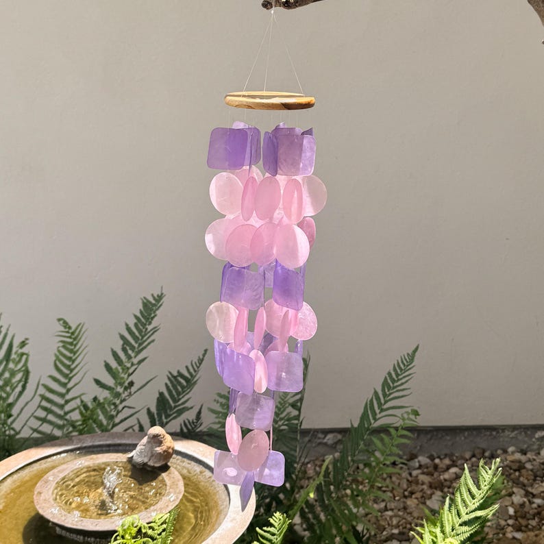 May include: A hanging wind chime with a wooden top and strands of pink and purple shell-like pieces. The wind chime is suspended from a small branch against a neutral background. The design is decorative and suitable for outdoor use.