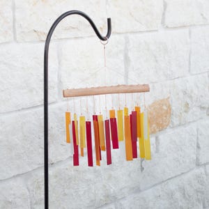 May include: A colorful wind chime with rectangular, translucent panels in shades of red, orange, and yellow. The panels hang from a wooden bar and are suspended by thin strings. The wind chime is mounted on a black metal hook against a white stone wall.