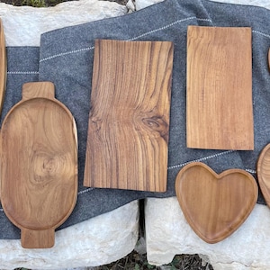 May include: A collection of seven wooden serving trays in various shapes, including rectangular, oval, leaf-shaped, and heart-shaped designs. Each tray features a natural light brown wood grain finish, showcasing the organic texture of the material.