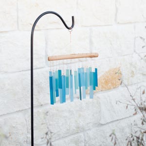 May include: A wind chime featuring multiple rectangular glass pieces in various shades of blue and teal, suspended by thin wires from a horizontal piece of light-colored driftwood. The chime hangs from a black metal shepherd's hook stand.