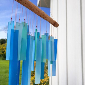 May include: A wind chime featuring multiple rectangular glass pieces in varying shades of blue, light blue, and teal. The glass rectangles are suspended by thin copper-colored wires from a horizontal piece of natural, textured brown driftwood.