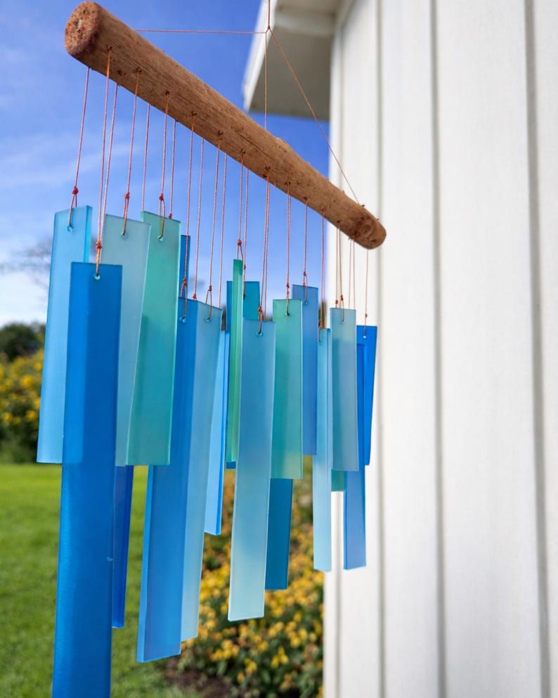 May include: A wind chime featuring multiple rectangular glass pieces in varying shades of blue, light blue, and teal. The glass rectangles are suspended by thin copper-colored wires from a horizontal piece of natural, textured brown driftwood.