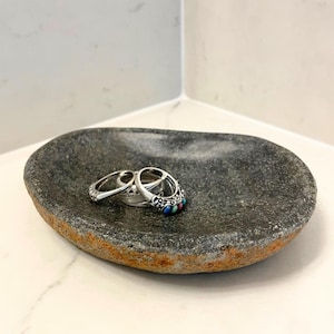 May include: A dark gray, oval-shaped stone dish with a rough, textured exterior and a smooth, concave interior. Resting inside the dish are two silver-toned rings, one featuring small, colorful inlaid stones in shades of blue, red, and green.