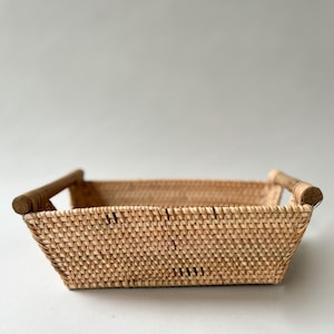 Peut inclure: Un panier rectangulaire tressé en fibres naturelles de couleur beige clair, présentant un motif de tissage serré et uniforme sur les côtés et la base. Le panier comprend deux poignées cylindriques en bois fixées aux côtés opposés.