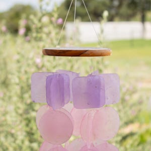 May include: A hanging wind chime with a wooden top and strings. The chime features square and round translucent pieces in shades of pink and purple. The background is a blurred view of greenery.
