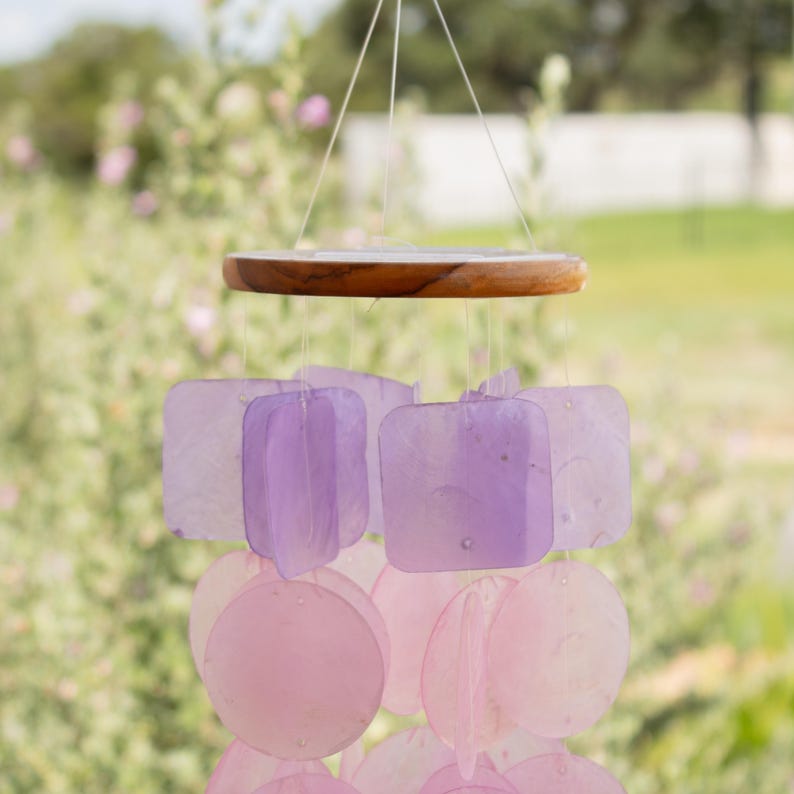 May include: A hanging wind chime with a wooden top and strings. The chime features square and round translucent pieces in shades of pink and purple. The background is a blurred view of greenery.