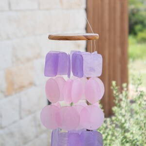 May include: A hanging wind chime with a wooden top and strands of iridescent, square and round, purple and pink shell pieces. The chime is suspended by thin, clear strings, and is set against a blurred outdoor background.