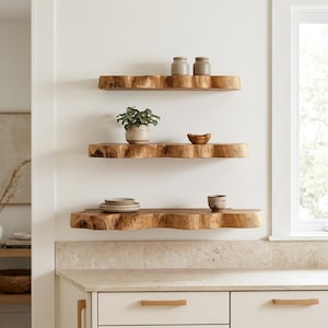 May include: Three wall-mounted floating shelves made of light brown natural wood with irregular live edges. The shelves are arranged vertically and hold two small jars, a potted green plant, a wooden bowl, a stack of plates, and a small ceramic cup.