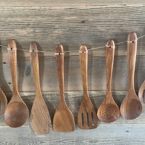 May include: A collection of eight wooden kitchen utensils, including spoons, spatulas, and a fork, are suspended from a string against a rustic wooden background. The utensils vary in shape and size, showcasing natural wood grain patterns and warm tones.