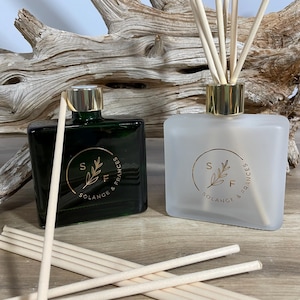 May include: Two glass diffuser bottles, one dark green and one frosted, with gold-colored caps and the text "SOLANGE & FRANCES". Each bottle has reeds inserted. Reeds are scattered in front of the bottles. The background includes driftwood.