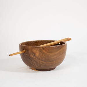 May include: A round, medium-brown wooden bowl with a visible natural grain pattern and a small circular hole on one side. A pair of light-colored wooden chopsticks is inserted through the hole, resting diagonally across the top of the bowl.