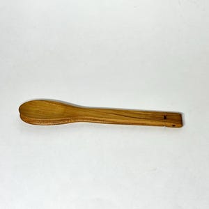 May include: A wooden spoon with a rounded bowl and a rectangular handle. The spoon is made of light brown wood and has a small hole at the end of the handle. The spoon is designed for cooking and serving.