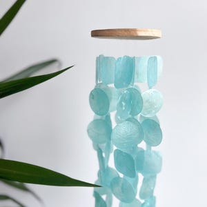 May include: A hanging mobile featuring a circular light-colored wooden top piece with multiple strands of translucent, light blue, flat, circular and square-shaped shell-like discs suspended from it, creating a layered, cascading vertical arrangement.