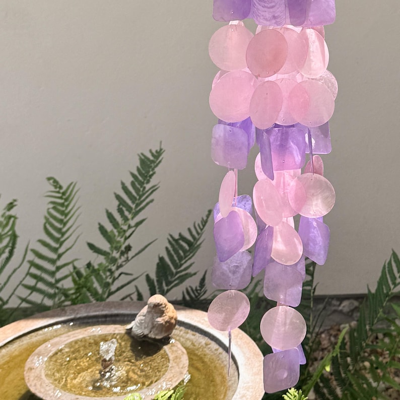 May include: A decorative wind chime with pink and purple shell-shaped pieces. The chimes are strung together, creating a cascading effect. The background includes a water fountain and greenery.