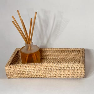May include: A rectangular woven rattan tray holds a glass diffuser bottle with wooden reeds. The bottle contains a golden-colored liquid. The tray and bottle are set against a white background, creating a neutral and natural aesthetic.