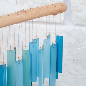 May include: A wind chime featuring a horizontal light brown wooden branch from which multiple rectangular glass pieces in varying shades of blue and teal are suspended by thin orange strings, creating a layered and vertical arrangement.