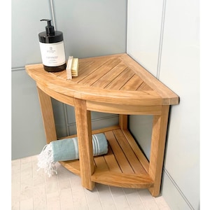 May include: A corner shower bench crafted from light brown teak wood, featuring a slatted top and a lower shelf. A black pump bottle labeled Cashmere and a small wooden brush sit on the top surface, while a folded teal towel rests below.