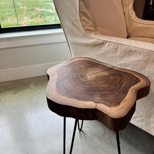 May include: A live edge wooden side table with a natural, organic shape. The tabletop is a rich brown color with lighter wood grain patterns. The table has three black metal hairpin legs, creating a modern, minimalist aesthetic.