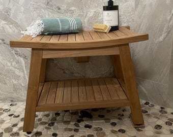 May include: A light brown wooden shower bench with a curved seat and a lower storage shelf. On the seat sits a folded sage green towel with white stripes and fringe, a small wooden brush, and a black pump bottle labeled Cashmere.