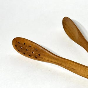 May include: Wooden kitchen tongs with a slotted head and a smooth, curved handle. The tongs are made of light brown wood and are designed for serving food. The image is taken against a white background.