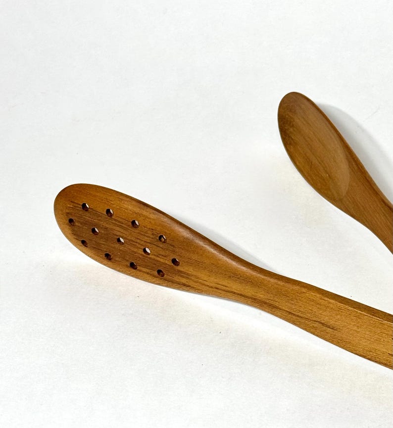 May include: Wooden kitchen tongs with a slotted head and a smooth, curved handle. The tongs are made of light brown wood and are designed for serving food. The image is taken against a white background.