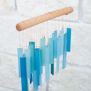 May include: A hanging wind chime featuring a horizontal light brown wooden branch from which multiple rectangular glass pieces in varying shades of blue and teal are suspended by thin copper-colored wires, creating a layered gradient effect.