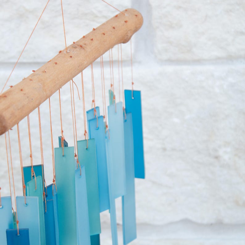 May include: A wind chime featuring a horizontal light brown wooden branch from which multiple rectangular glass pieces in various shades of blue are suspended by thin orange strings. The glass pieces vary in length and opacity, creating a gradient.