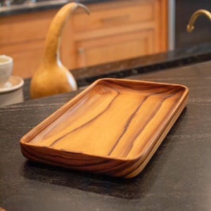 May include: A rectangular wooden serving tray with rounded corners. The tray is made of light brown wood with darker brown grain patterns. The tray is sitting on a dark countertop.