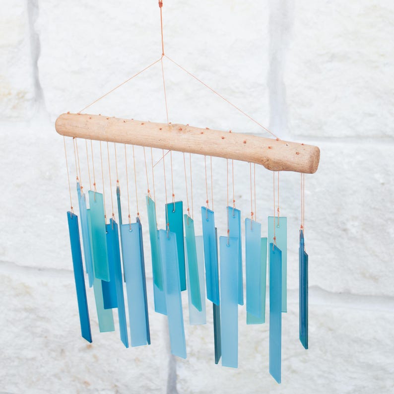 May include: A wind chime featuring a horizontal piece of light-colored driftwood suspended by thin copper wires. Numerous rectangular glass pieces in various shades of blue, ranging from pale aqua to deep navy, hang from the wood.