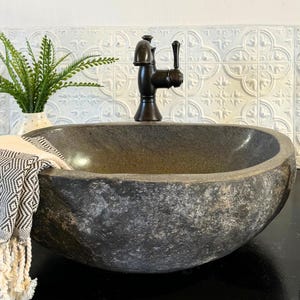 May include: A dark gray, natural stone vessel sink with a rough, textured exterior and a smooth, polished interior basin. A dark bronze-toned metal faucet is mounted behind the sink. A black and white patterned textile with fringe hangs over the side.